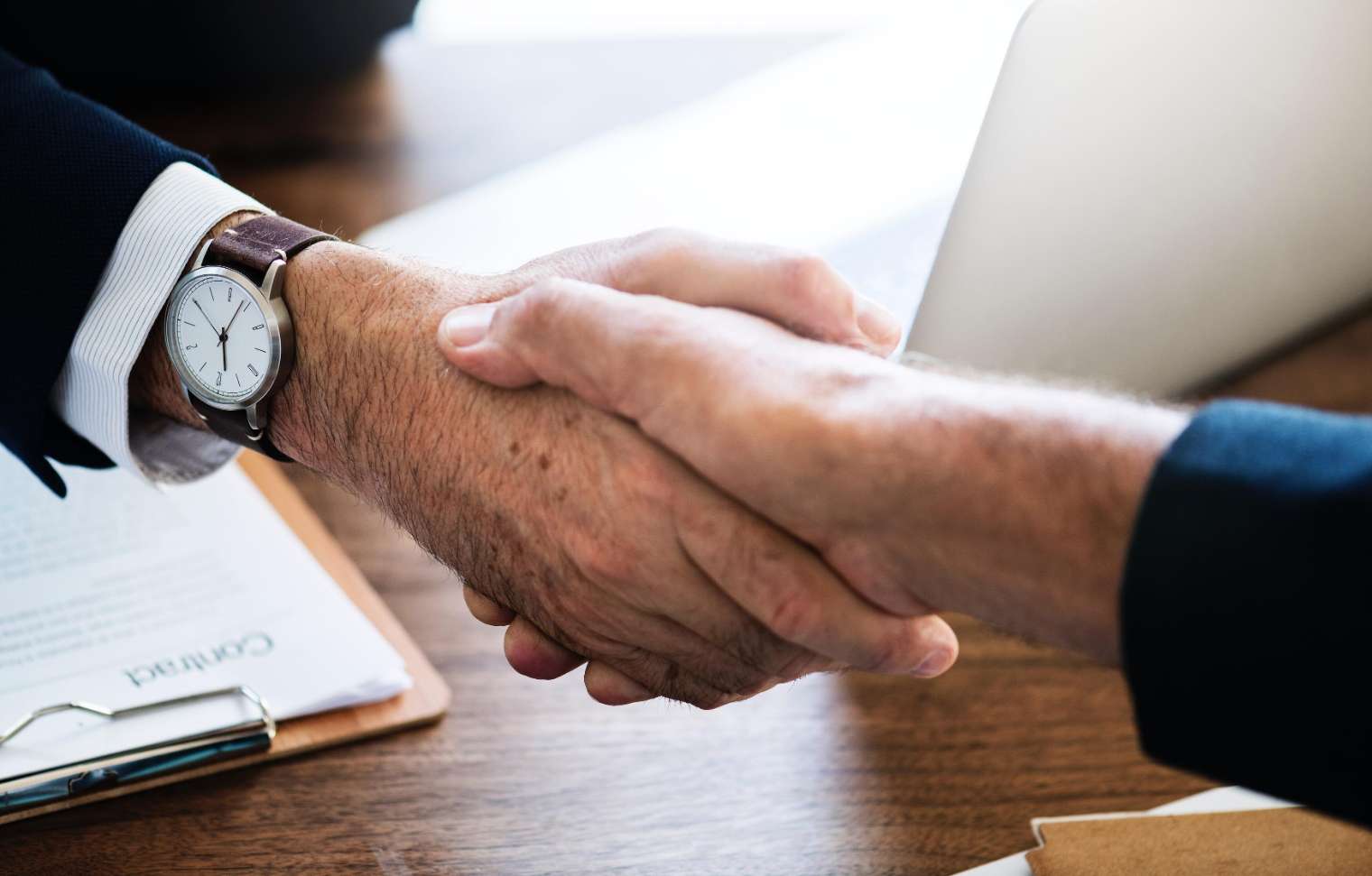 two men shaking hands over desk with open laptop and clipboard